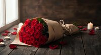 Romantic red roses bouquet wrapped in kraft paper on rustic wooden table with candles and scattered petals creating intimate atmosphere for special occasions.