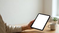 Person holding tablet with blank screen at desk with coffee cup and small plant, suitable for app mockups, technology presentations, and digital marketing materials.
