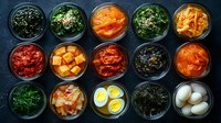 Korean side dishes in glass bowls including kimchi, seaweed, pickled vegetables, marinated eggs and fermented accompaniments on dark background, traditional banchan.