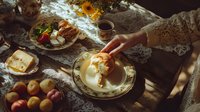 Female hands in lace sleeves serving traditional breakfast with fresh bread, butter, fruits and coffee on rustic wooden table in morning sunlight through window.