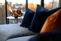 Cozy evening atmosphere with navy blue velvet pillows on grey sofa, decorative candle holder with warm lights against window view during golden hour sunset.
