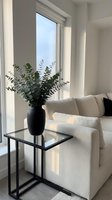 Modern minimalist living room interior with eucalyptus branches in black vase on glass side table next to white sofa by large window with natural light.