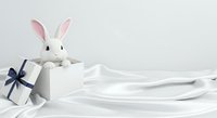 White rabbit toy with pink inner ears peeking from gift box with navy bow on white silk fabric background, suitable for Easter, holiday and celebration designs.