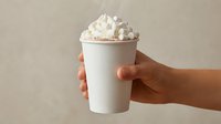 Hand holding a steaming white paper cup topped with whipped cream and marshmallows, perfect for winter beverage and cafe menu concepts.