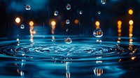 Water drops falling into calm blue water creating concentric ripples, with glowing golden bokeh lights in background against dark surface. Macro photography captures liquid motion.