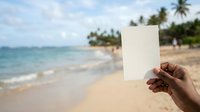 Unrecognizable person holding blank white card on tropical beach with turquoise water, palm trees, and sandy shore, suitable for travel and vacation marketing.