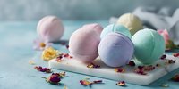 Colorful bath bombs in pastel pink, purple, and mint green on marble tray with dried flower petals against turquoise background for spa and self-care.