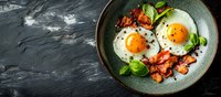 Sunny side up eggs with crispy bacon strips and fresh basil leaves served on green ceramic plate against dark stone background, panoramic banner format.