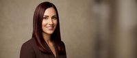 Professional businesswoman with long brown hair smiling confidently in dark blazer against neutral background for corporate and career content.