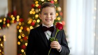 Young Caucasian boy in formal black tuxedo with red bow tie holding single red rose, smiling at camera with Christmas tree bokeh lights background.