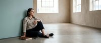 Woman sitting alone on floor in empty room with natural light from windows, suitable for mental health, loneliness, and wellness concepts.