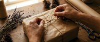 Hands wrapping kraft paper gift box with twine and dried lavender flowers on wooden table for eco-friendly present packaging.