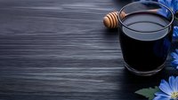 Glass of black activated charcoal latte with honey dipper and blue flowers on dark wooden background, modern healthy detox drink for wellness.