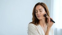 Young Caucasian woman applying makeup with brush in bright bathroom, morning beauty routine skincare cosmetics preparation for daily wellness lifestyle.