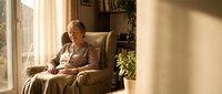 Elderly Caucasian woman relaxing in comfortable armchair by sunny window in cozy living room with warm natural lighting and home interior decor.