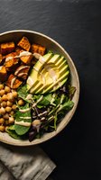 Healthy vegan Buddha bowl with roasted sweet potato, sliced avocado, chickpeas and mixed greens on dark slate background for nutritious meal planning.