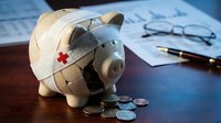 Piggy bank with bandage and red cross surrounded by coins on desk with financial documents and pen representing healthcare savings and medical expenses.