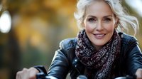 Mature woman with blonde hair wearing leather jacket and knit scarf smiling outdoors in autumn park with natural bokeh background and warm lighting.