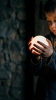 Young Asian woman fortune teller holding glowing crystal ball in dark mystical setting with warm light illuminating her face and hands creating magical atmosphere.