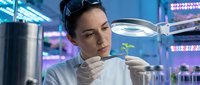 Female scientist examining plant seedling under magnifying glass in modern laboratory with LED grow lights for botanical research and agricultural development.