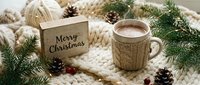 Cozy Christmas flat lay with hot cocoa mug, Merry Christmas wooden sign, pine cones, and fir branches on chunky knit blanket.