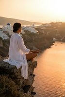 Young woman practicing meditation on cliff overlooking coastal village at golden hour sunset, wearing white flowing clothes in peaceful mindfulness pose.