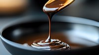 Liquid chocolate or caramel sauce pouring from spoon into dark bowl creating smooth glossy swirl, macro food photography with selective focus and blur background.