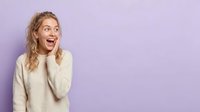 Young woman with wavy blonde hair wearing cream sweater expresses surprise and excitement against purple background, suitable for lifestyle and emotion-based designs.