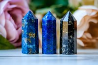 Three polished crystal towers in blue lapis lazuli and black obsidian arranged on white surface with soft floral background for healing and meditation practices.
