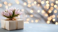White gift box with pink flower decoration and gold ribbon against blurred bokeh lights background creating festive holiday atmosphere.