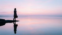 Woman silhouette walking on calm water surface during sunset with pink purple sky reflection creating peaceful meditation scene for wellness concepts.