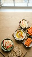 Healthy snack spread with hummus, apple slices, carrot sticks and crackers on wooden table by window for nutritious eating and meal prep.