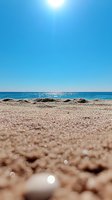 Bright sun shining over calm blue sea with sandy beach in foreground, creating sparkling reflections on water surface and sand grains.