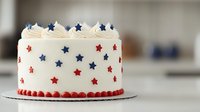 Patriotic white frosted cake decorated with red and blue stars and pearl border, topped with whipped cream and star accents for holiday celebration.