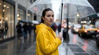 Woman in bright yellow raincoat holding transparent umbrella on rainy urban street with blurred city background and traffic. Caucasian female, casual style, wet weather conditions.