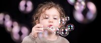 Young curly-haired child blowing soap bubbles against dark background with floating iridescent spheres creating magical childhood moment for family content.