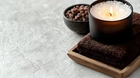 Glowing aromatic candle on brown towel with coffee beans in bowl on gray concrete background, spa wellness composition for relaxation and aromatherapy.