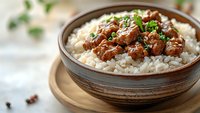 Tender braised beef chunks in rich brown sauce served over steamed white rice in rustic ceramic bowl, garnished with fresh herbs and black pepper.