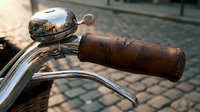 Vintage motorcycle handlebar with weathered brown leather grip and chrome bell reflecting sunlight, parked on cobblestone street for retro transport and nostalgia themes.
