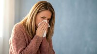 Woman with brown hair and light skin tone blowing nose into tissue while wearing pink sweater, depicting cold, flu, or allergy symptoms for healthcare and wellness content.