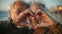 Senior couple making heart shape with hands at sunset beach, romantic elderly love gesture, golden hour lighting for Valentine's Day cards and relationship concepts.