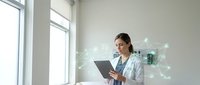 Young Asian female doctor in white coat using digital tablet with holographic medical network interface in modern hospital office for telemedicine consultation.