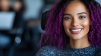 Young mixed race woman with vibrant purple curly hair and bright smile wearing gray sweater against blurred background, radiating confidence and joy.