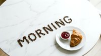 Word MORNING spelled with coffee beans on marble table beside croissant and berry jam on white plate, cozy breakfast concept.