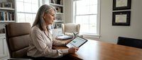 Professional blonde businesswoman using tablet computer at wooden desk in modern home office with natural lighting and bookshelves.