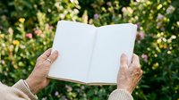 Senior adult hands holding open blank notebook against lush green foliage background, suitable for education, publishing, and lifestyle content.