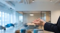 Businessman in dark suit gesturing with open palm in modern office with glass partitions and blurred colleagues, conveying communication, presentation, or agreement concept.