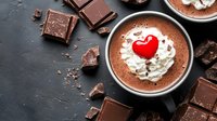 Hot chocolate topped with whipped cream and red heart decoration served in dark mug, surrounded by chocolate pieces on black background. Perfect for Valentine's Day.