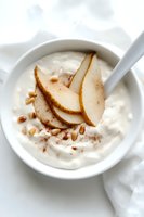Fresh sliced pears with pine nuts and cinnamon served over creamy white yogurt in ceramic bowl, high-key food photography on light background.