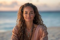 Woman with long wavy brown hair and Mediterranean ethnicity wearing pink linen shirt with pendant necklace, smiling at beach during golden hour sunset.